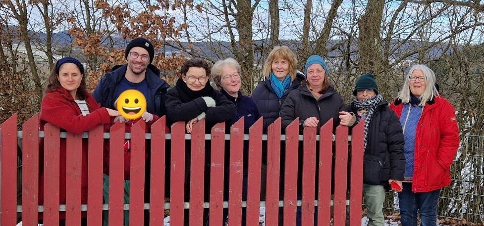 Neun Menschen stehen nebeneinander an einem Gartenzaun und lächeln in die Kamera.